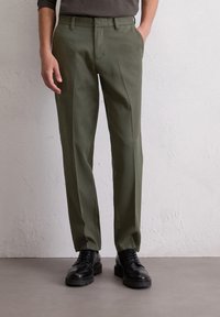 Pantalons vert olive foncé en tissu lisse, avec une taille plate, des poches latérales et une coupe ajustée, associés à des chaussures noires.