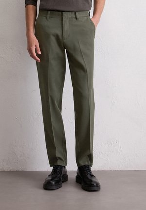 Donker olijfgroene broek van een gladde stof, met een platte voorkant, zijzakken en een getailleerde pasvorm, gecombineerd met zwarte schoenen.
