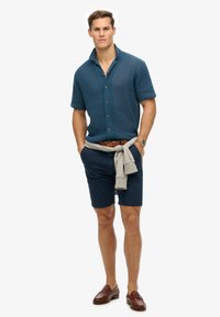 Kortærmet, tekstureret mørkeblå skjorte med krave, beige knapper; marineblå shorts; lys grå sweater bundet om taljen; brune læder loafers.