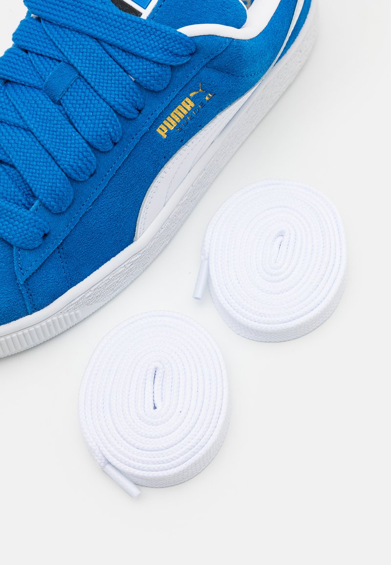 Baskets en suède bleu avec des accents blancs, dotées de lacets épais. Deux lacets ronds blancs supplémentaires sont placés à côté de la chaussure sur une surface blanche.