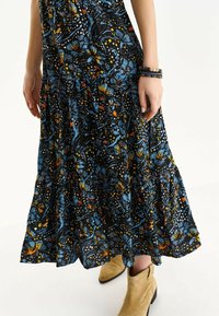 Vestido largo negro con estampados vibrantes en azul, naranja y blanco. Doquier acampanado, tejido ligero, combinado con botines beig.