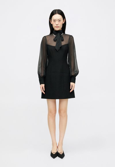Robe noire avec une silhouette ajustée, des manches longues transparentes et un décolleté en nœud papillon. La texture du tissu est lisse avec un léger drapé.