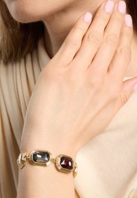 Pulsera de oro con tres piedras octagonales: gris, roja y transparente. Montada en un diseño de eslabones ondulados de oro sobre un fondo de tela clara.