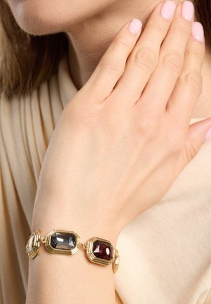 Goldarmband mit drei achteckigen Edelsteinen: grau, rot und klar. Eingefasst in ein wellenförmiges Goldverbindungsdesign vor einem hellen Stoffhintergrund.