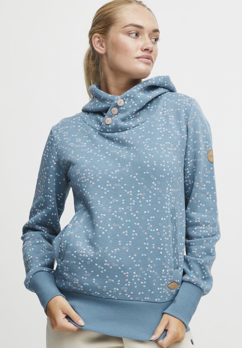 Sudadera azul claro con cuello tipo cowl, tres botones y un patrón de pequeños círculos en rosa y blanco. Incluye detalles de puños y dobladillo acanalados.