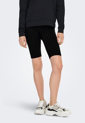 Personne portant un short de cycliste noir, un haut à manches longues noir et des baskets blanches et grises épaisses, debout sur un fond uni.