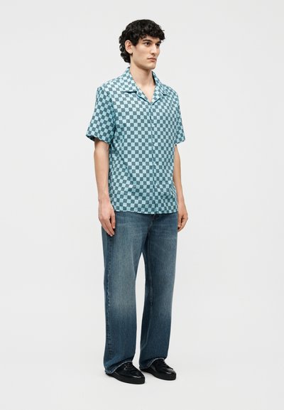 Άνδρας φορώντας μπλε καρό κοντομάνικο πουκάμισο, φαρδύ μπλε τζιν και μαύρα παπούτσια, στέκεται μπροστά σε απλό ανοιχτό φόντο.
