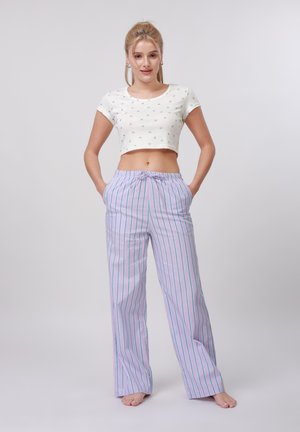 Femme debout, pieds nus, portant un t-shirt blanc court avec de petits imprimés et un pantalon de pyjama rayé lavande taille haute, les mains dans les poches.