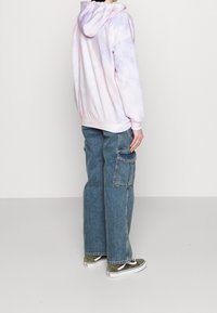 Jasnoróżowy i fioletowy hoodie tie-dye z kapturem, w połączeniu z luźnymi niebieskimi spodniami cargo z dżinsu, które mają boczne kieszenie i mankiety na końcach.