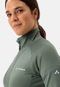 Veste verte à col haut faite de matériau texturé, dotée d'une fermeture éclair à l'avant et d'un logo subtil sur la poitrine et l'épaule.