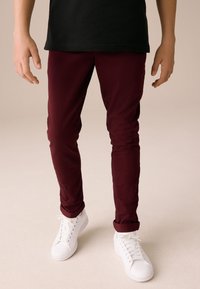 Pantalon stretch bordeaux à coupe slim, avec revers roulés, porté avec des baskets blanches et un t-shirt noir. Texture lisse, design décontracté.