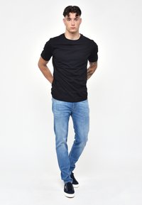 T-shirt nera a maniche corte con scollatura rotonda, abbinata a jeans di denim azzurri e sneakers blu navy. Il modello mostra uno stile casual e aderente.