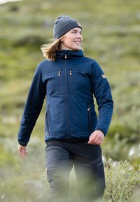Marineblaue Softshelljacke mit einem Reißverschluss vorne und Brusttaschen, kombiniert mit schwarzen Hosen, getragen mit einer grauen Mütze. Freiluftumgebung.