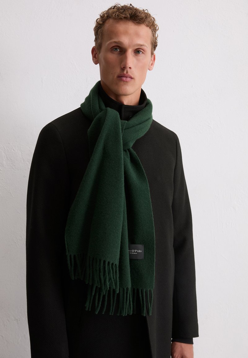 Sciarpa verde scuro realizzata in tessuto morbido, con dettagli frangiati alle estremità e un piccolo etichetta con il logo. Abbinata a un cappotto nero.