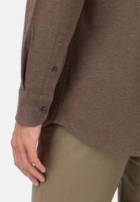 Hnědá košile s dlouhým rukávem vyrobená z texturované látky, s černými knoflíky na manžetách, mírně zakřiveným lemem a volným střihem.