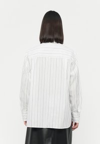 Camicia bianca con bottoni, a righe nere sottili, maniche lunghe e vestibilità rilassata. La parte posteriore presenta un design a yoke e un dettaglio a tab vicino al colletto.