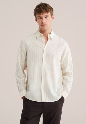 Bărbat tânăr, în picioare, cu mâinile în buzunare, purtând o cămașă crem cu mâneci lungi, cu nasturi și dungi verticale subțiri, și pantaloni închiși la culoare.