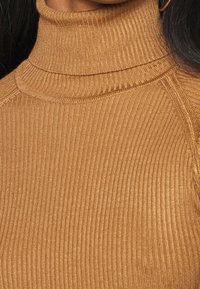 Zbliżenie osoby noszącej prążkowany, wielbłądzi golf i złotą kolczyk koło, z widocznymi długimi ciemnymi włosami.