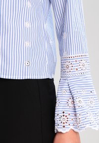 Blusa a righe blu e bianche con accenti di pizzo floreale ricamato sulle maniche a campana. Il tessuto sembra leggero, con una vestibilità leggermente strutturata.
