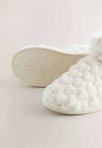 Chaussons blancs et moelleux avec des motifs de cœur en relief sur le dessus et des semelles à motifs d’étoiles sur un fond clair uni.