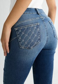 Jeans in denim blu scuro con due tasche posteriori, una delle quali presenta un motivo a rombi e piccoli borchie. Fit su misura con una finitura leggermente sbiadita.