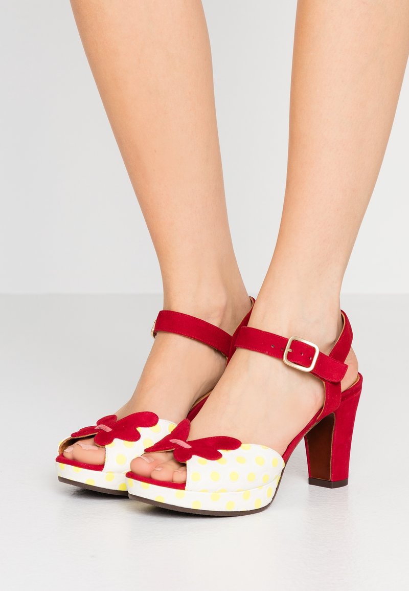 Pieds portant des sandales rouges à talons hauts avec des brides à la cheville, présentant des plateformes blanches à pois jaunes et des décorations rouges en forme de papillon.
