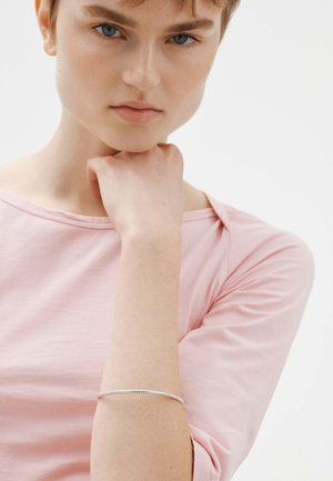Joven con el cabello corto, vestido con una camiseta rosa claro, descansando la barbilla en la mano, llevando una delgada pulsera de plata en la muñeca, frente a un fondo liso.