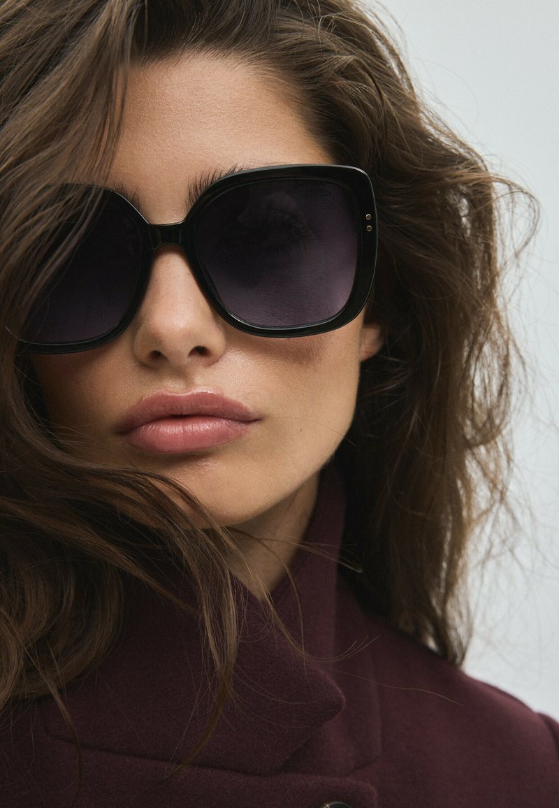 Femme aux longs cheveux bruns portant de grandes lunettes de soleil noires et un manteau violet foncé, regardant légèrement sur le côté sur un fond clair.