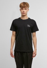 Black cotton t-shirt with short sleeves, round neckline, featuring white graphic text on the left chest area, displayed on a neutral backdrop.