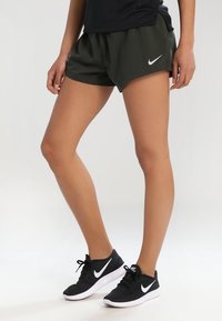 Musta Nike juoksukengät, tummanvihreät urheilushortsit, kevyt materiaali, löysä istuvuus, ikoninen Nike-vipu-logo shortsissa ja kengissä.