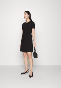 Svart kortermet kjole med tettsittende bodice og utsvingt skjørt, laget av glatt stoff. Båret med svarte slingback-sko og en liten håndveske.