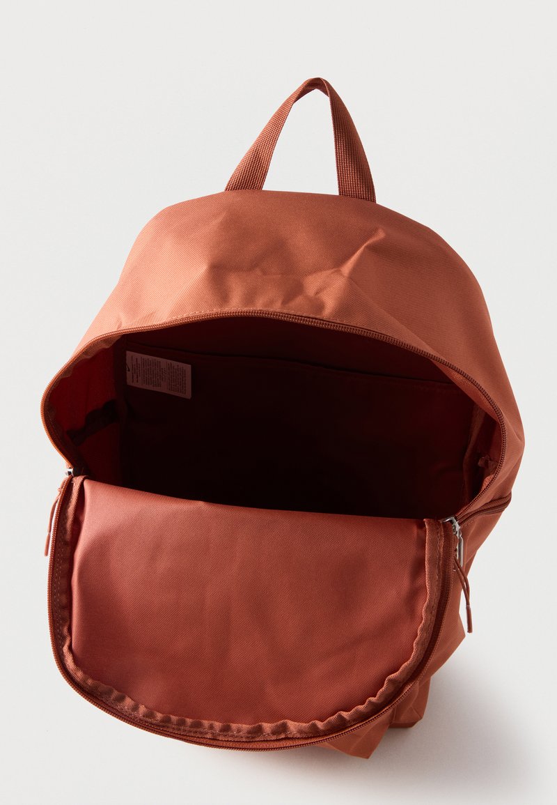 Mochila abierta de color óxido con un gran compartimento principal con cremallera y una asa de tela en la parte superior, mostrada sobre un fondo claro y liso.