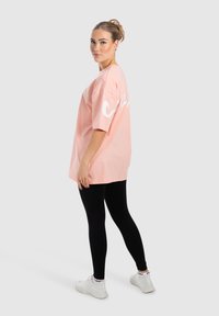 Lyserød, oversized t-shirt i bomuld med hvid grafisk tekst på ryggen, kombineret med sorte leggings og hvide sneakers. Glat struktur, afslappet pasform.