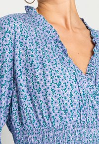 Květinové šaty v světle fialové barvě s zelenými a modrými akcenty. Mají výstřih do V, volány a nabíraný pas pro texturu.