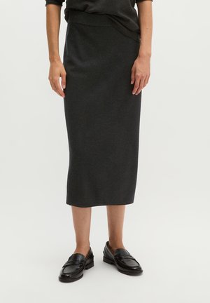 Femme portant une jupe crayon midi gris foncé et des mocassins en cuir noir, debout devant un fond clair uni.