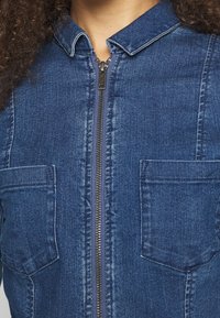 Jeansowy kombinezon z suwakiem z przodu, kołnierzem i dwoma kieszeniami na piersi. Charakteryzuje się głębokim niebieskim kolorem oraz teksturowaną tkaniną z widocznymi szwami.