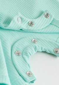 Tissu côtelé vert menthe avec boutons-pression argentés partiellement fermés sur un vêtement de bébé sur fond blanc.