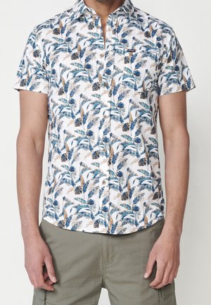 Homme portant une chemise à manches courtes boutonnée avec un motif de feuilles tropicales bleu et marron et un pantalon cargo vert olive.