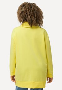 Sweatshirt jaune surdimensionné avec un col montant, des manches longues et une coupe décontractée. Texture lisse avec une ligne d'ourlet atteignant les hanches.