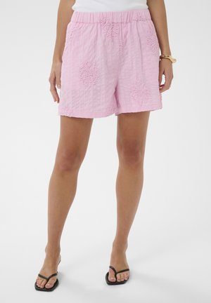 Donna che indossa pantaloncini rosa testurizzati con motivi floreali, top bianco, sandali neri e un braccialetto d'oro al polso destro.