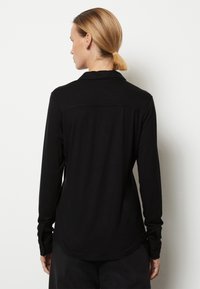 Camisa negra de manga larga hecha de un tejido suave. Presenta un cuello clásico, puños abotonados y un dobladillo curvado. Simple, sin patrones visibles.