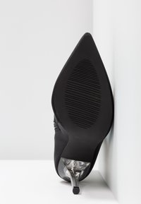 Chaussure à talon haut noire à bout pointu vue par la semelle avec un talon aiguille transparent reposant contre un mur blanc.