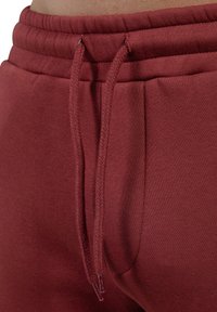 Pantalones de chándal color marrón con una cintura elástica y cordón ajustable. Textura de tela suave, con bolsillos laterales y detalles de costura sutiles.