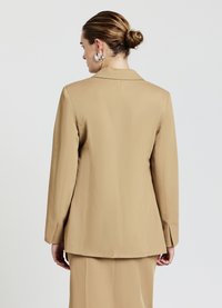 Blazer tan con maniche lunghe, vestibilità sartoriale e revers a ribalta. Tessuto liscio e una fessura sul retro, abbinato a una gonna coordinata.
