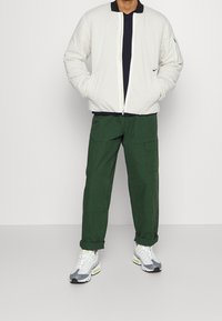 Light-colored puffer jacket with zip, navy t-shirt, green cargo pants, and gray/white sneakers. Notable zip pocket on jacket sleeve.