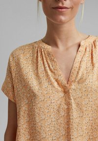 Kurzärmlige Bluse aus leichtem orangefarbenem Stoff mit floralem Muster. Sie hat einen Ausschnitt mit Schlitz und geraffte Details an den Schultern.