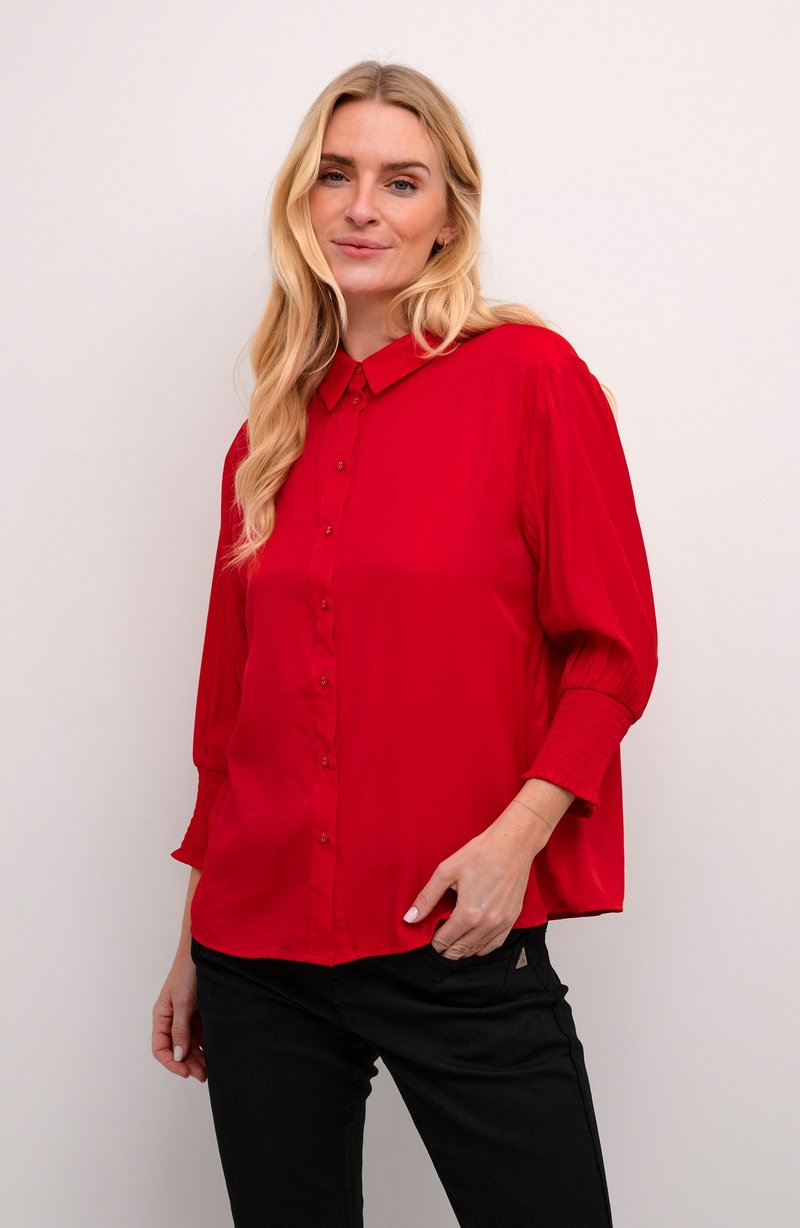 Punane särk nööpidega, kraega, 3/4-sleeve'id elastsete mansettidega. Sile kangas ja mugav lõige, koos mustade pükstega.
