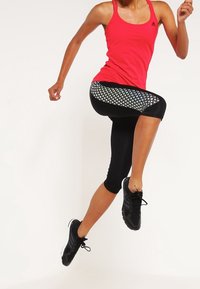 Rød atletisk tanktop med tynde stropper, parret med sorte capri leggings med et netmønstret panel. Sorte atletiske sko.