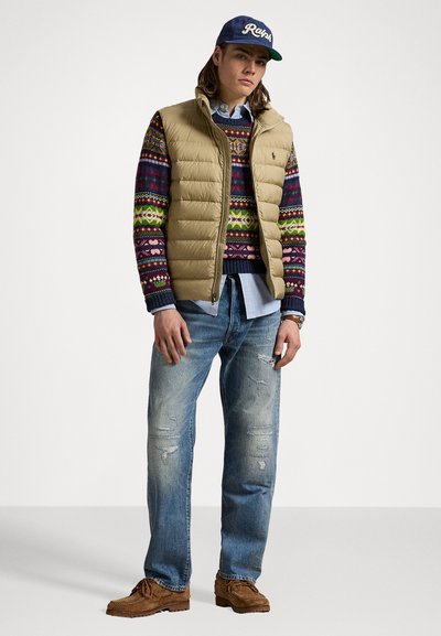 Beige puffer vest over a multicolored patterned sweater, light blue shirt, distressed blue jeans, and brown moccasin shoes.