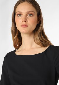 Schwarzes Kleid mit einem breiten, quadratischen Ausschnitt und weichem, strukturiertem Stoff. Das Model hat lange, wellige Haare und einen natürlichen Make-up-Look.